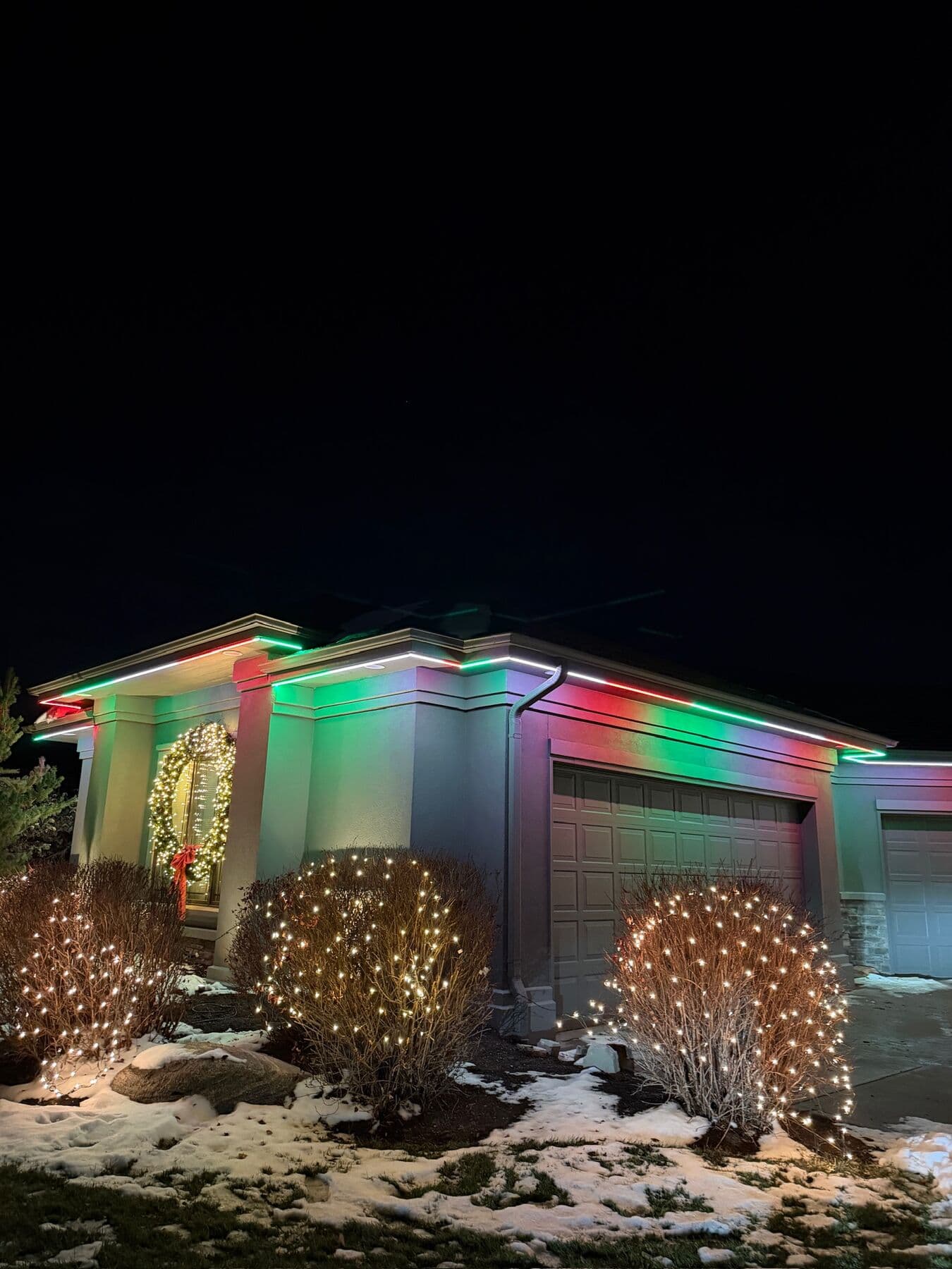 A stucco Omaha entry in green and red with three round shrubs under net lights and snow on the beds