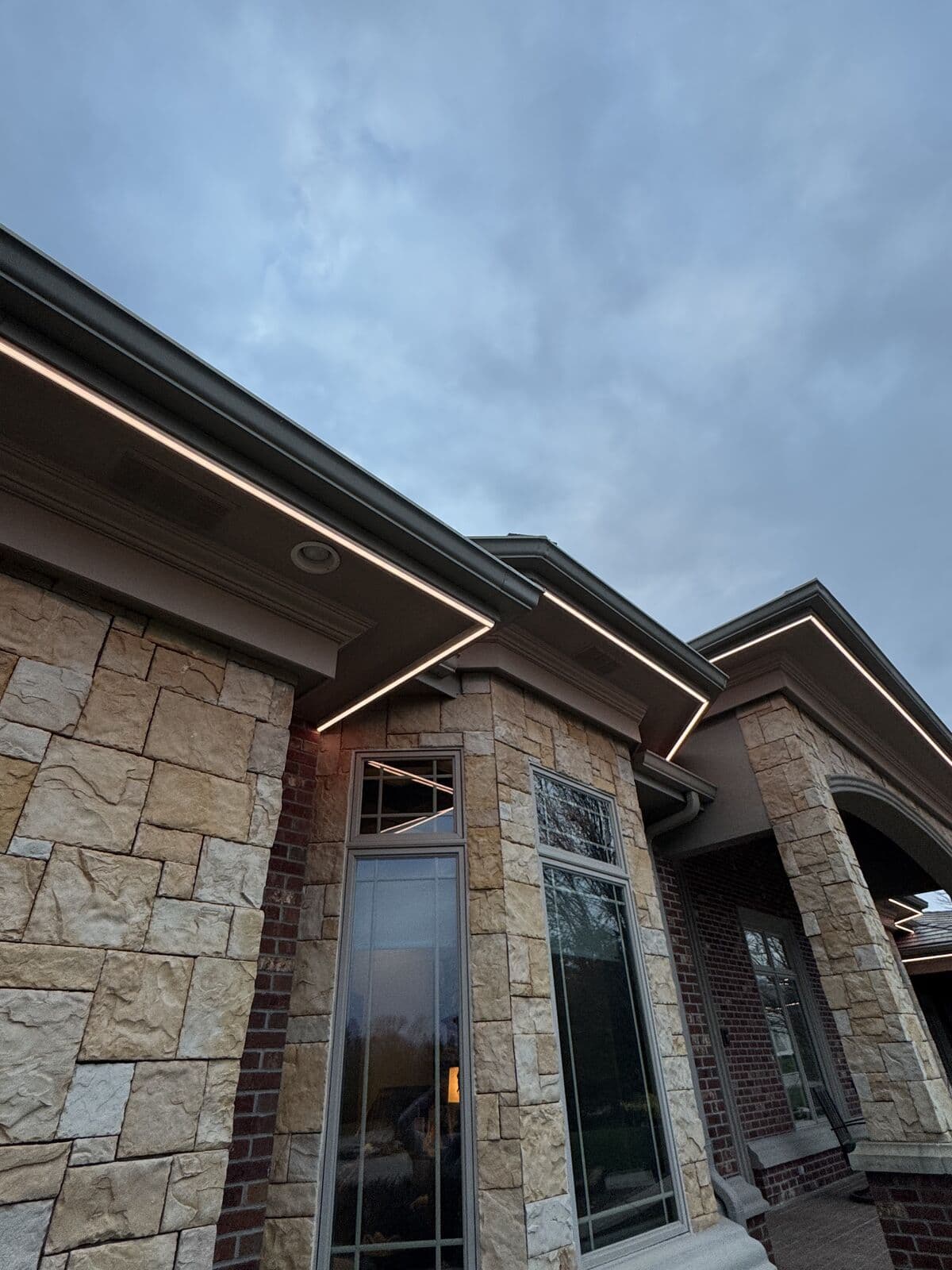 A stone and brick gable on an Omaha home under overcast light, the channel following the roof edge above the entry