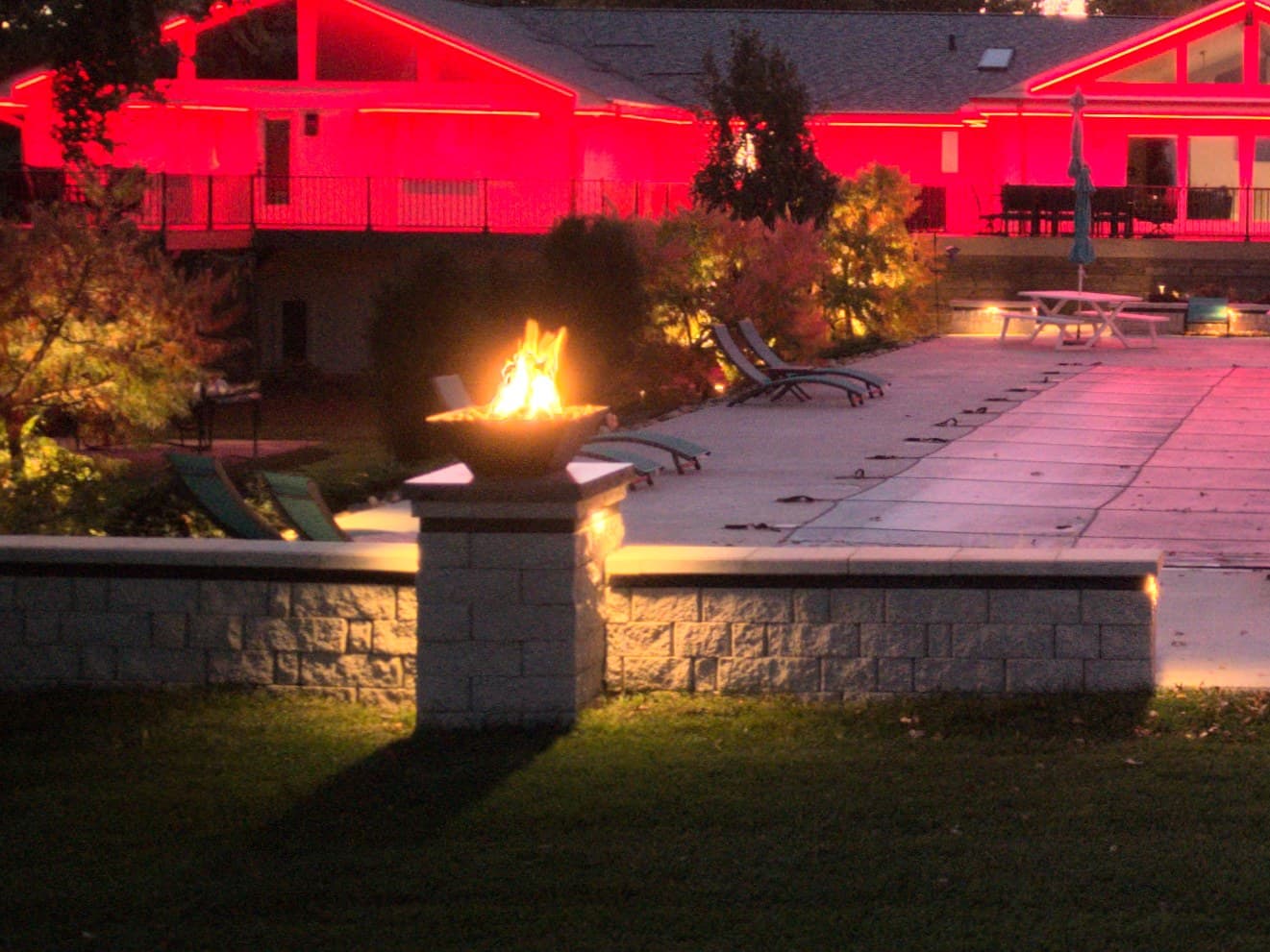 An Omaha back garden at night: a lit seat wall, a fire bowl on a pier, and the house roofline behind set to red