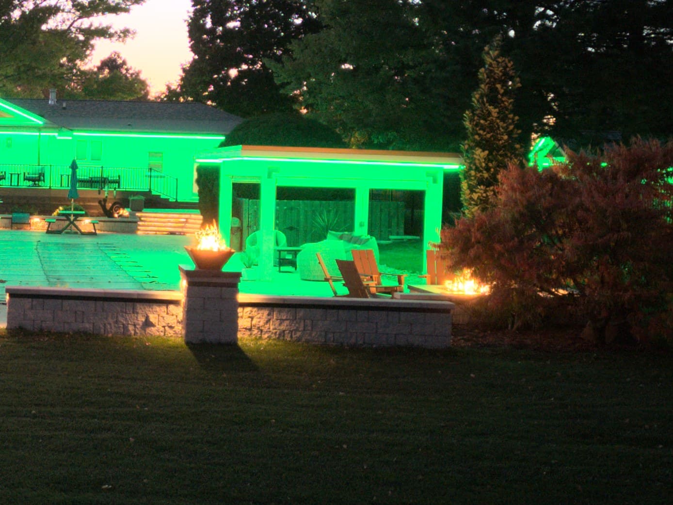 A lit pergola over a pool patio at an Omaha home, the roofline and pergola fascia set to green, with a fire bowl on a seat wall