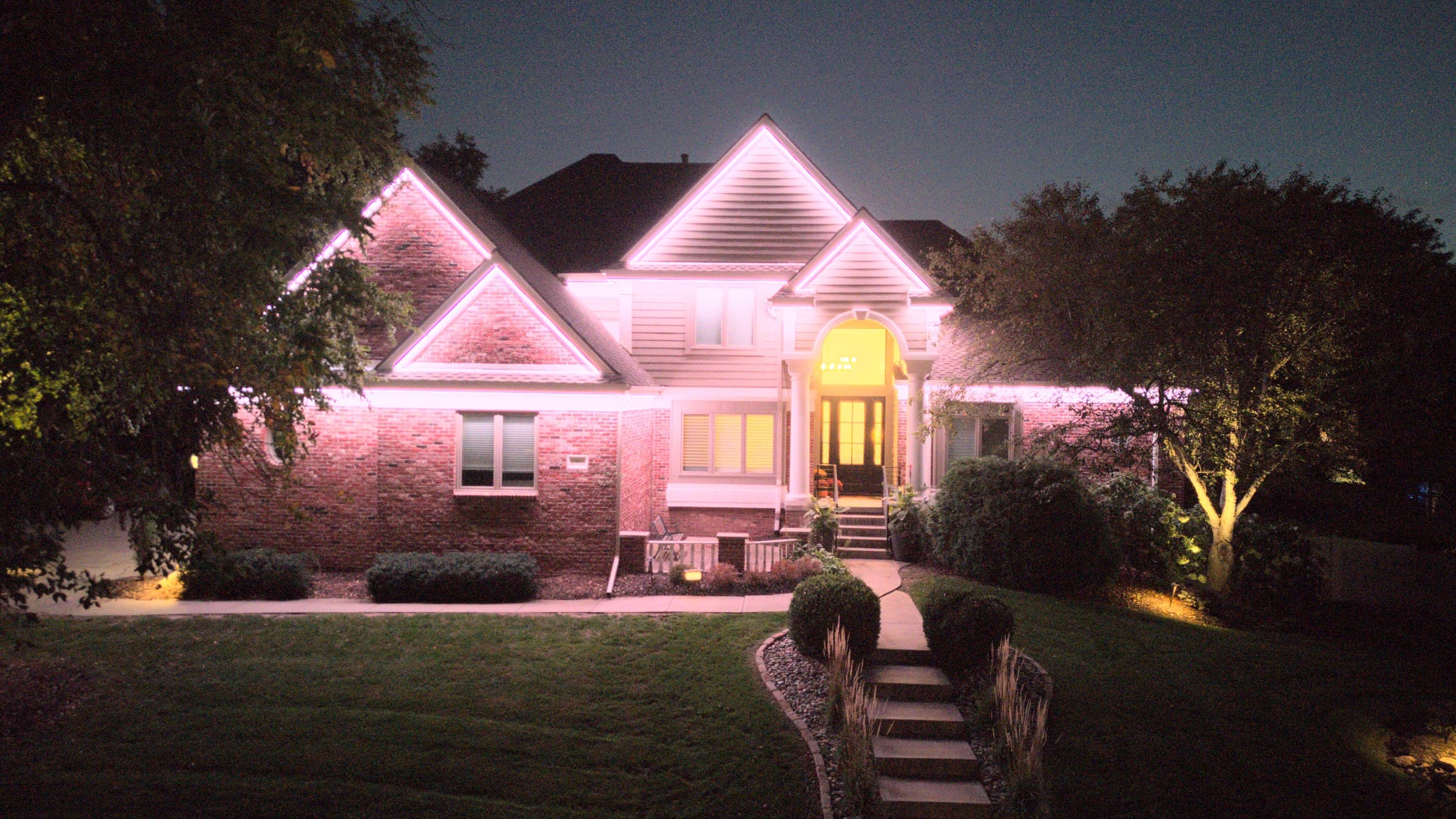 An Omaha two-story with every roofline run on the same soft pink, and the front walk and beds lit as well as the roofline