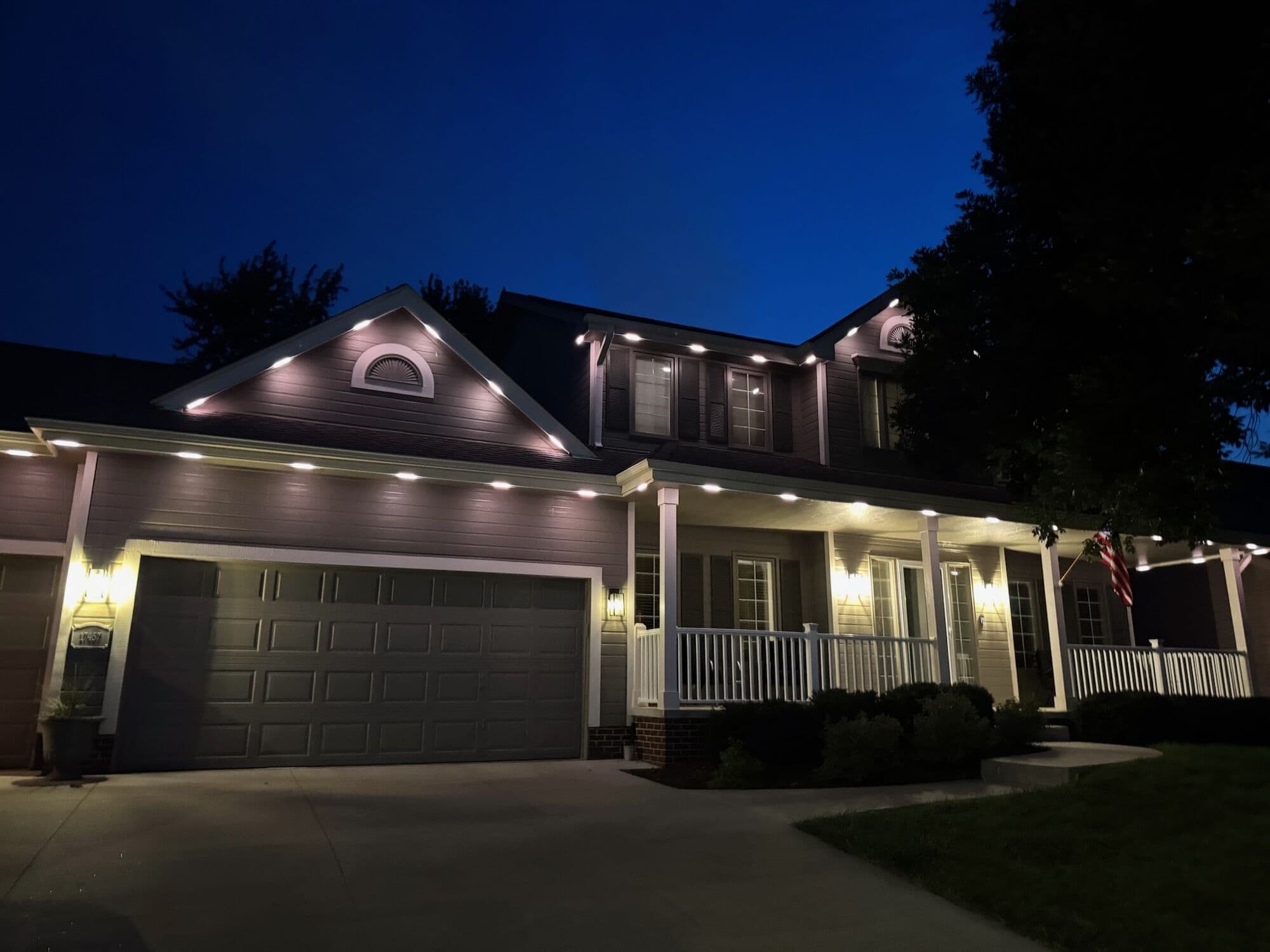 A gray Omaha two-story at blue hour with warm downlights over the porch and a flag by the door