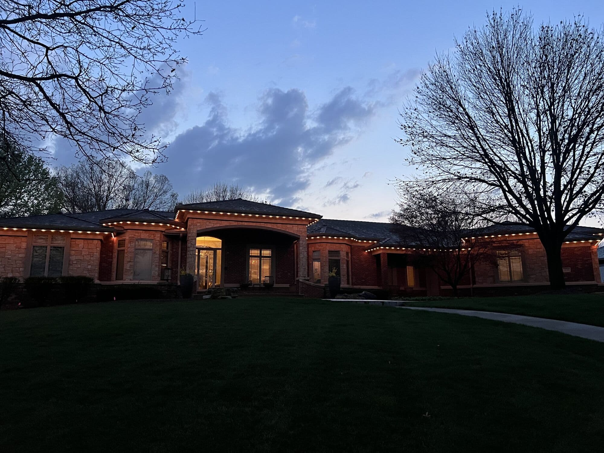 A long Omaha ranch at blue hour behind a bare tree, warm white along the whole roofline