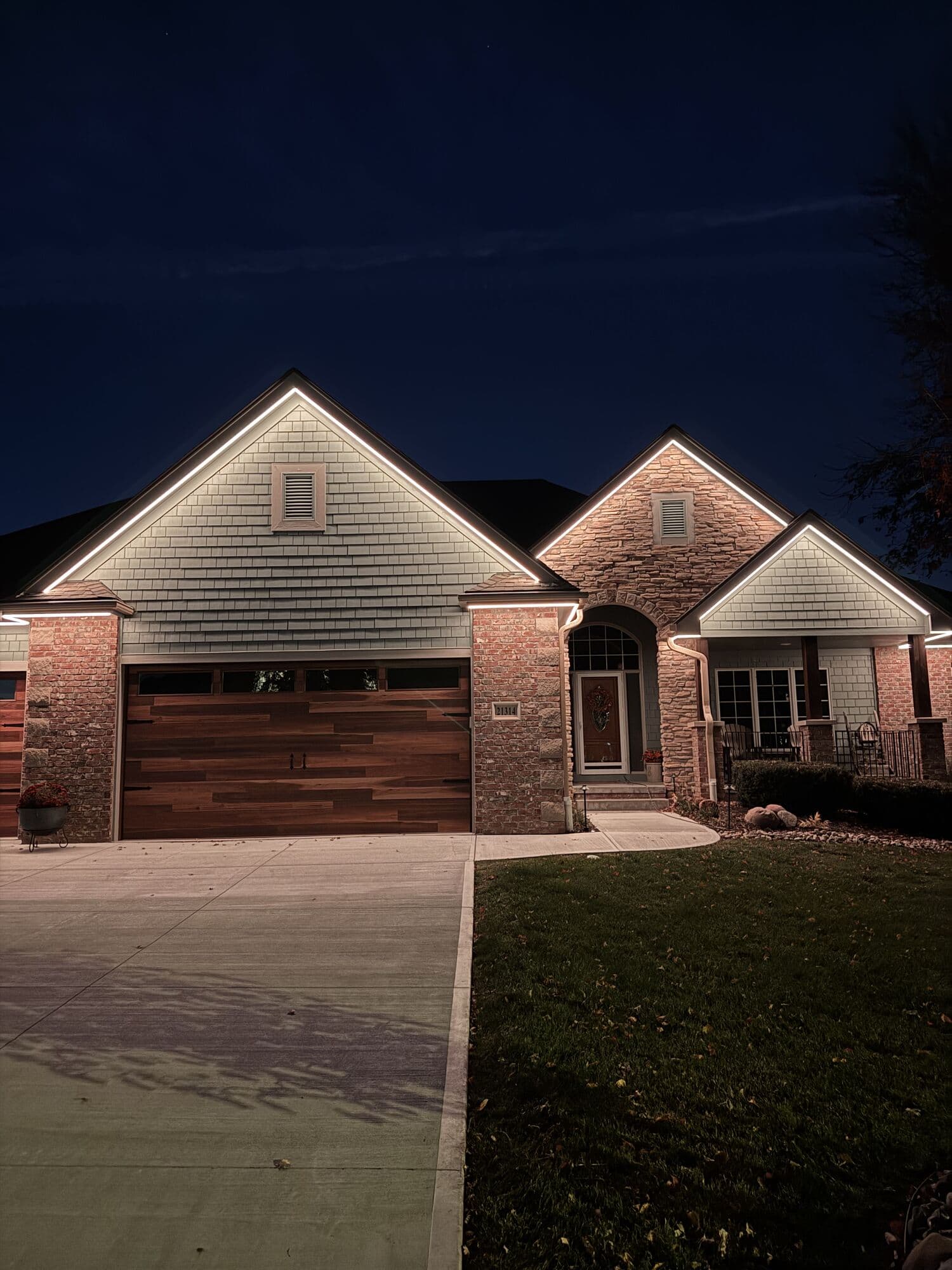 An Omaha home in shake and brick with a wood garage door, warm white downlights along every eave and gable
