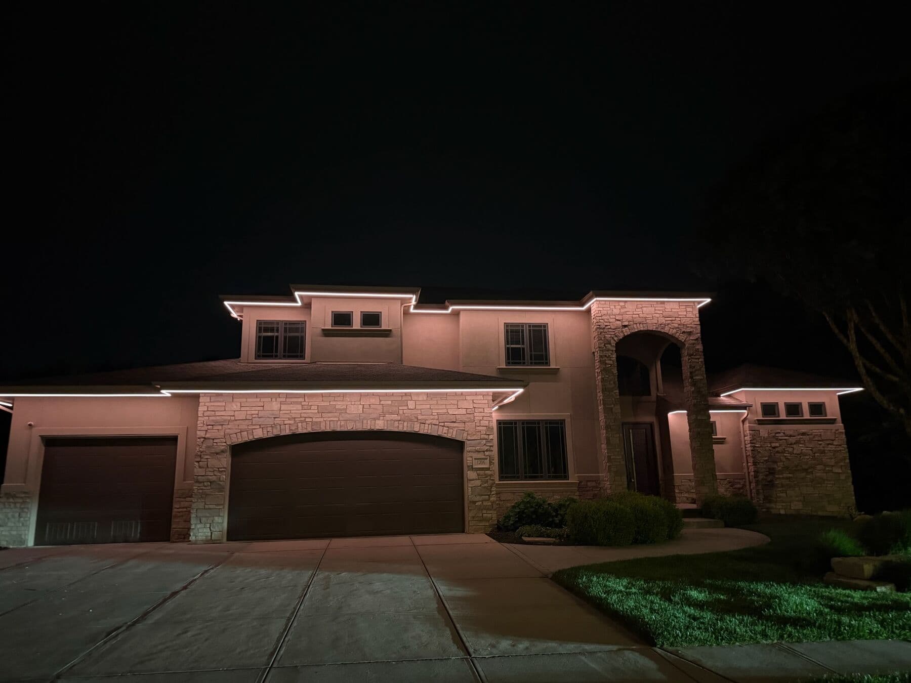 A stucco and stone Omaha two-story lit warm along the rooflines with a green uplight in the bed below