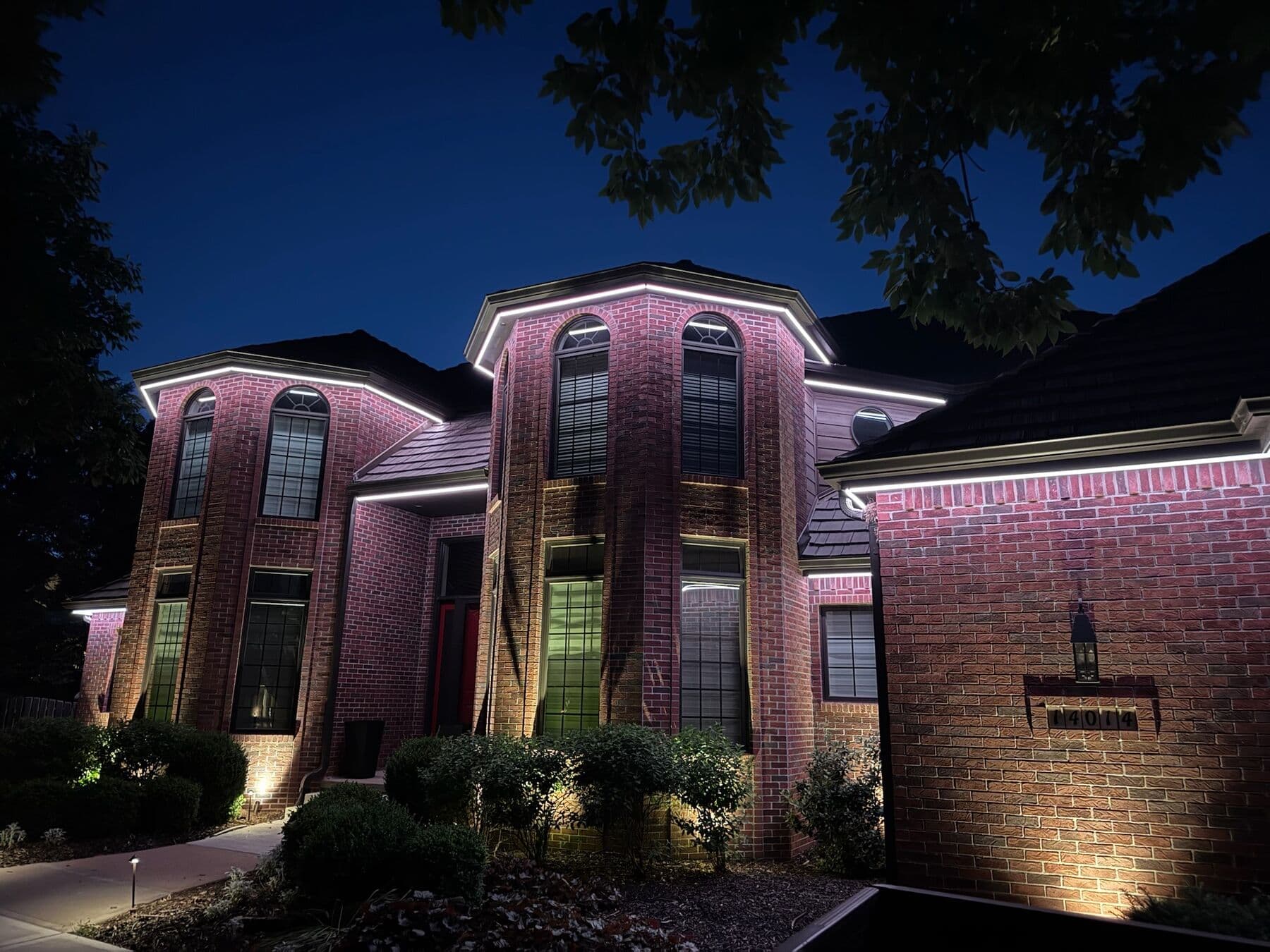 A tall brick Omaha home with white roofline runs, magenta accents on the gables and uplights washing the brick and shrubs