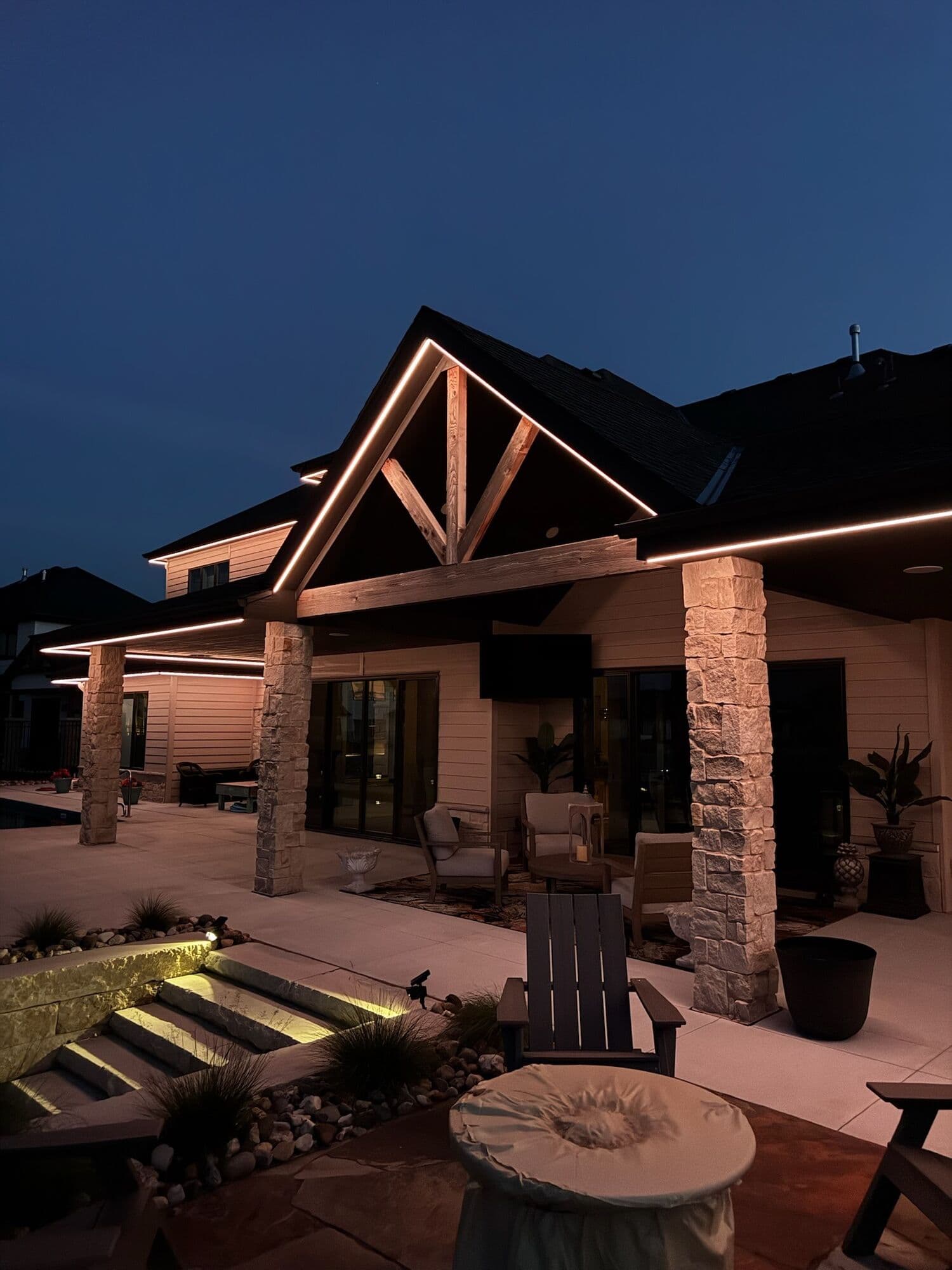 A timber and stone covered patio at an Omaha home after dark, warm light along the beam and step lights down the stairs