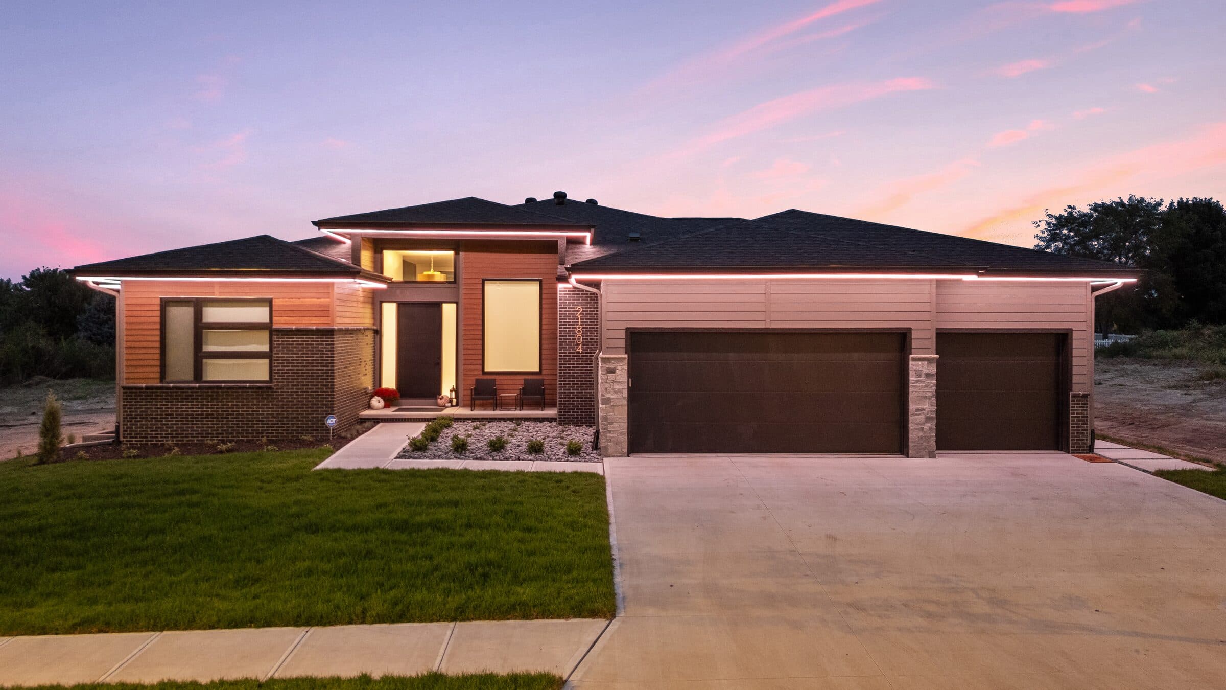 A brick and cedar ranch west of Omaha at dusk, its roofline, eaves and garage band picked out in everyday warm white