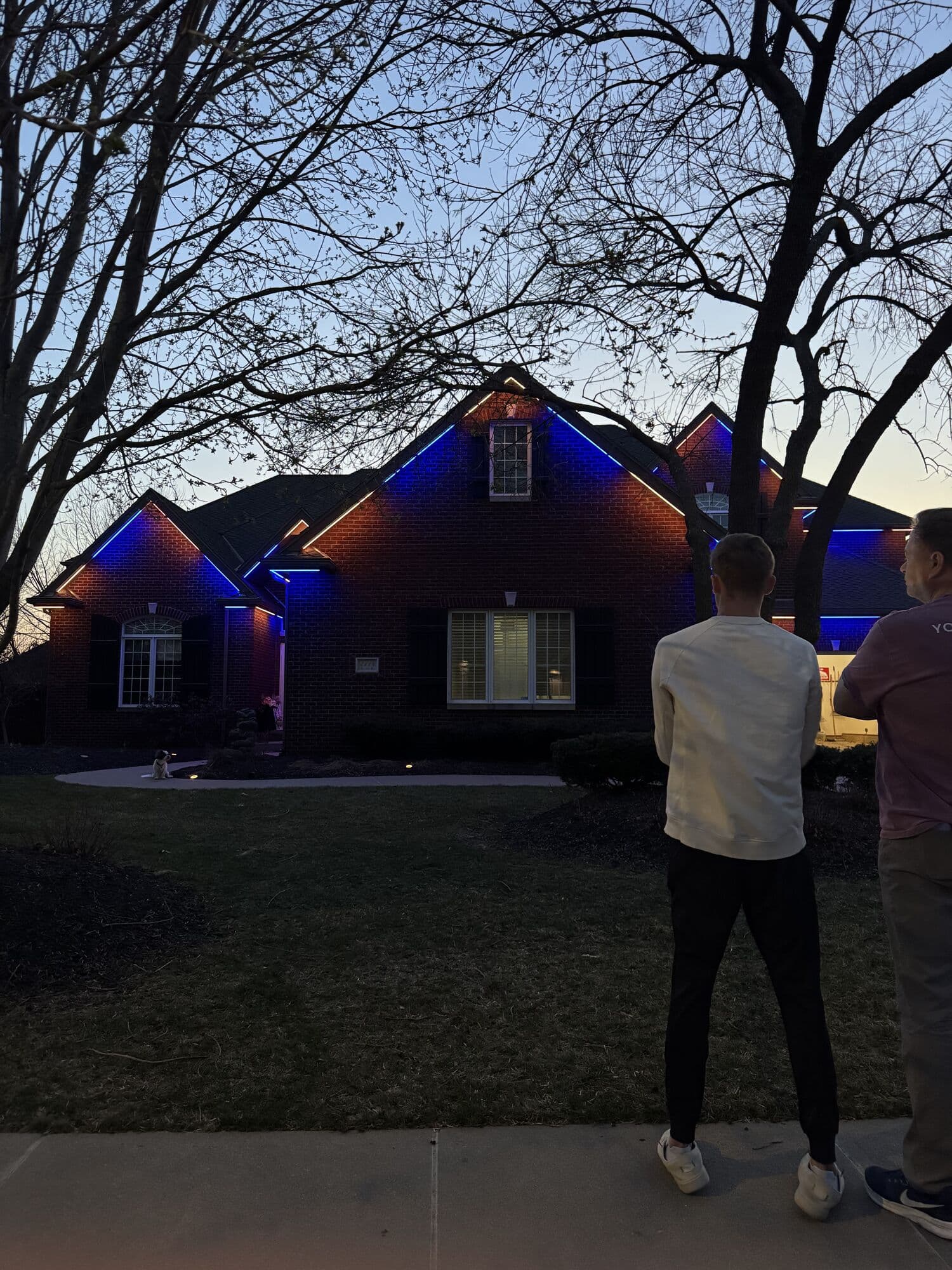 Two people standing on a lawn at dusk looking up at an Omaha home lit in blue and red
