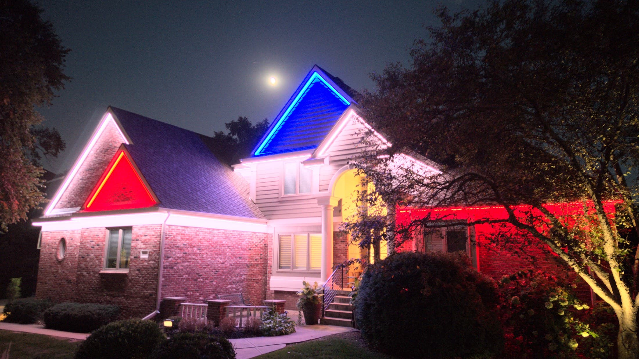 A two-story Omaha home with its roofline, gables and front trees lit at night